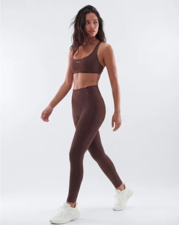 A woman modeling a matching brown activewear set consisting of a sports bra and high-waisted leggings, paired with white sneakers, against a light gray studio background.