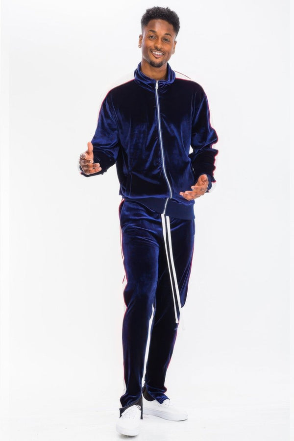 A man modeling a matching navy blue velvet tracksuit with white piping details, featuring a zip-up jacket and matching pants, paired with white sneakers against a plain white background.