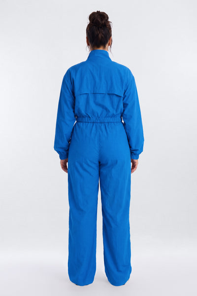 A person viewed from the back wearing a bright blue one-piece jumpsuit or coverall with long sleeves, a high collar, and wide legs, standing against a plain light gray studio background.