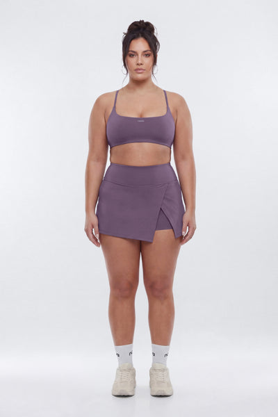 A woman modeling a matching athletic set consisting of a solid purple sports bra and wrap-style mini skirt,