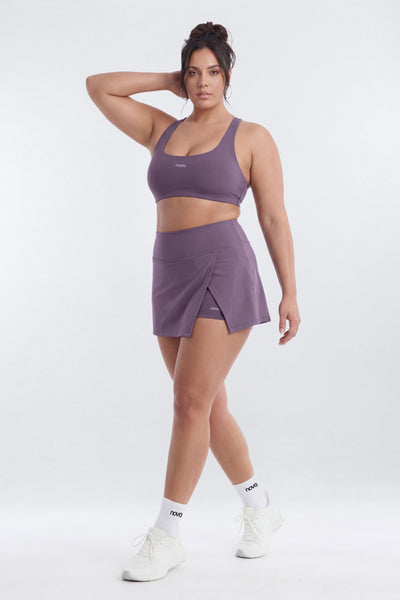 A woman modeling a matching athletic set consisting of a purple sports bra and a wrap-style mini skirt,