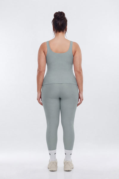 A woman viewed from behind wearing a matching sage green active wear set consisting of a tank top and leggings against a plain white studio background.