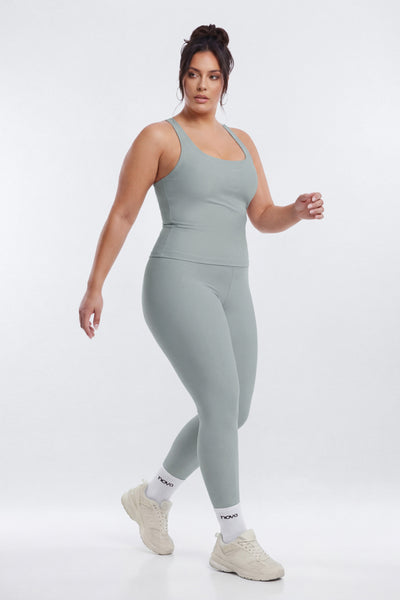 A woman modeling a matching sage green active wear set consisting of a tank top and leggings against a light gray studio background.