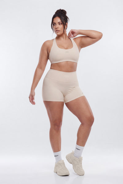 A woman modeling a matching beige activewear set consisting of a sports bra and high-waisted shorts, paired with white socks and beige sneakers, against a light gray studio background.