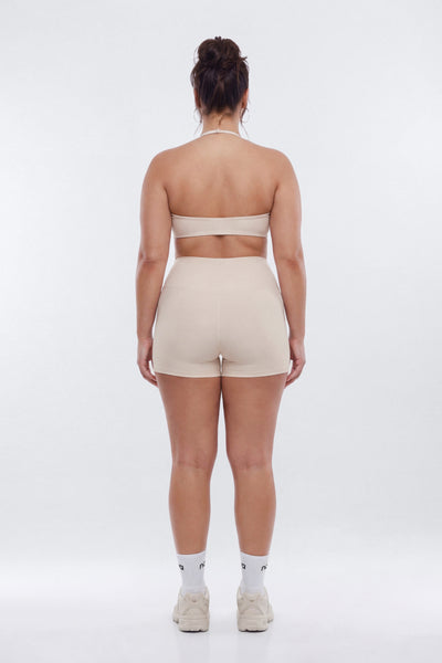 A woman viewed from behind wearing a matching beige two-piece activewear set consisting of a halter-style sports bra and high-waisted shorts, paired with white socks and beige sneakers on a light gray studio background.
