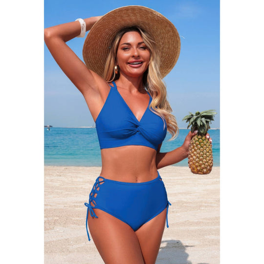 A woman on a beach wearing a bright blue two-piece bikini and a large straw hat, holding a pineapple. The background shows the blue ocean and sky.