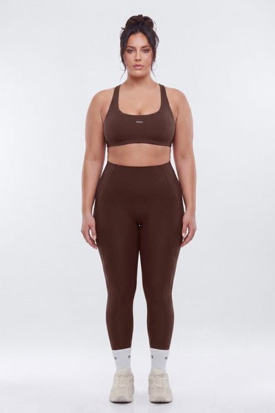 A woman modeling a matching brown activewear set consisting of a sports bra and high-waisted leggings, paired with white socks and beige sneakers, against a light gray studio background.