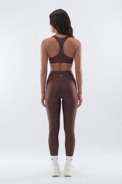 A woman viewed from the back wearing a matching dark brown activewear set consisting of a sports bra and high-waisted leggings, paired with white socks and sneakers, against a light gray studio background.
