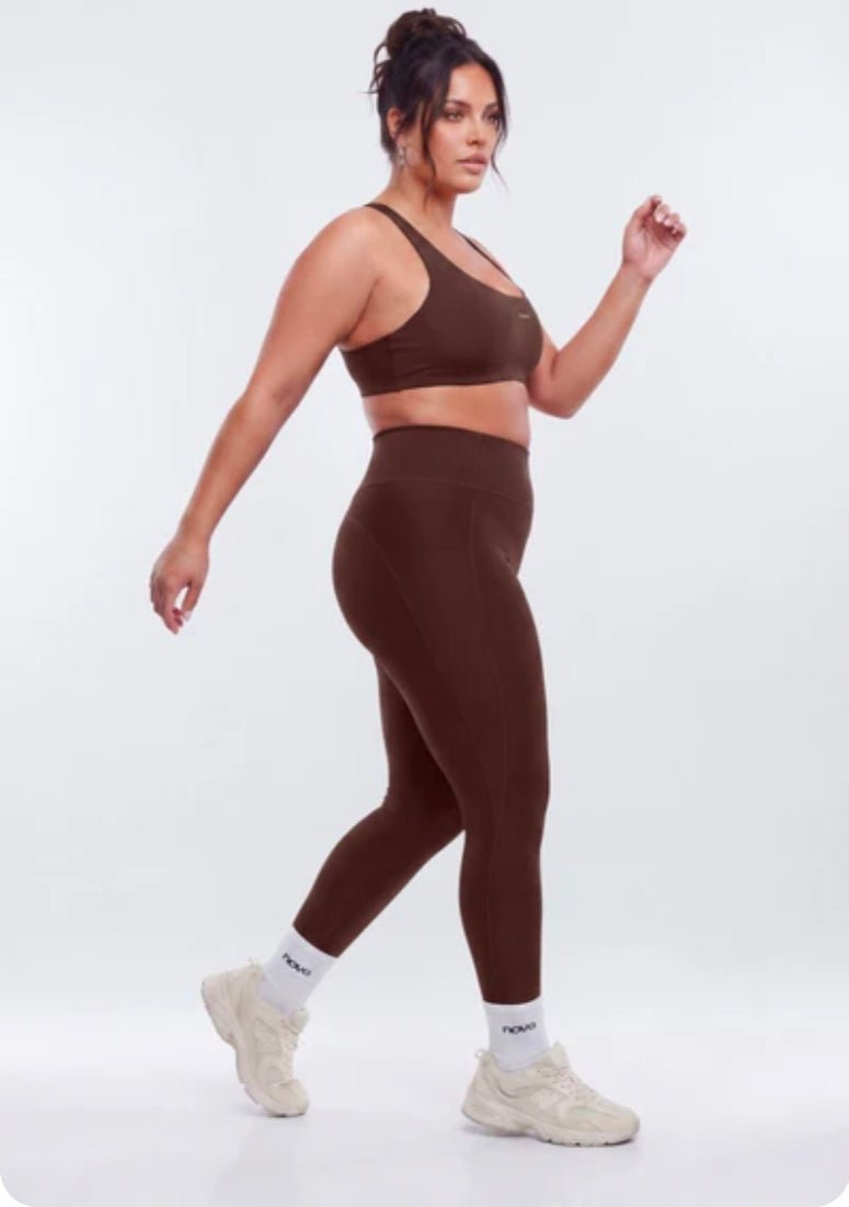 A woman modeling a matching brown activewear set consisting of a sports bra and high-waisted leggings, paired with white socks and beige sneakers, against a light gray studio background.