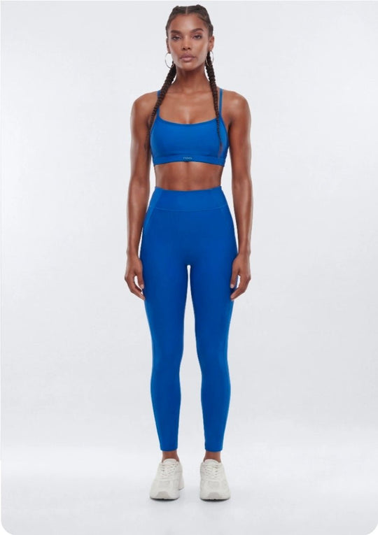 A woman modeling a matching athletic set consisting of a bright blue sports bra and high-waisted leggings, paired with white sneakers, against a light gray studio background.