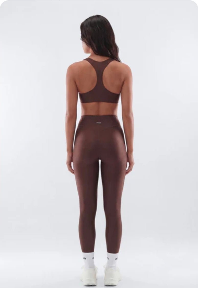 A woman viewed from the back wearing a matching dark brown activewear set consisting of a sports bra and high-waisted leggings, paired with white socks and sneakers, against a light gray studio background.