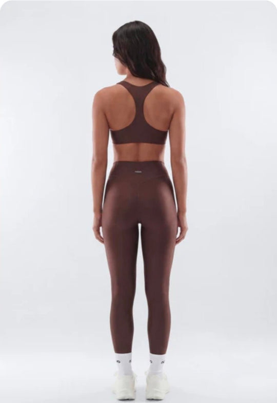 A woman viewed from the back wearing a matching dark brown activewear set consisting of a sports bra and high-waisted leggings, paired with white socks and sneakers, against a light gray studio background.