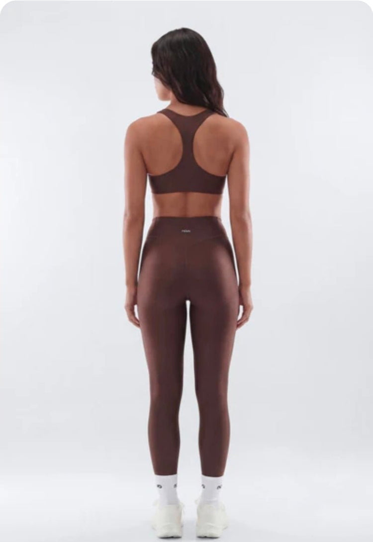 A woman viewed from the back wearing a matching dark brown activewear set consisting of a sports bra and high-waisted leggings, paired with white socks and sneakers, against a light gray studio background.