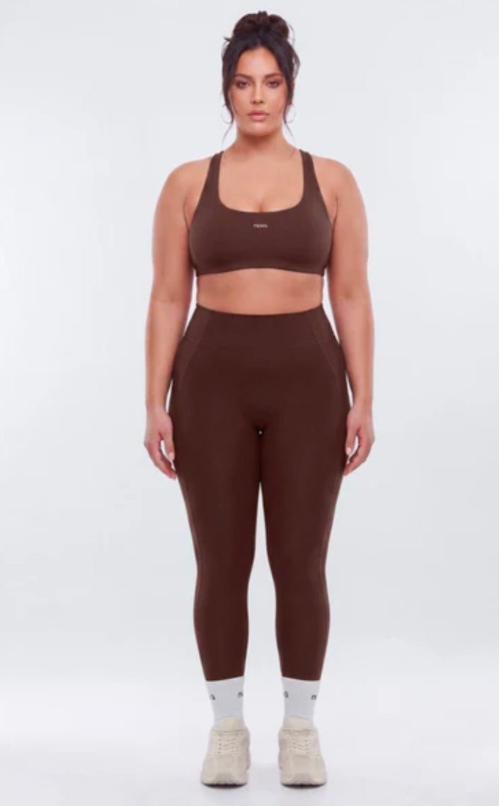 A plus-size model stands against a light gray studio background wearing a matching brown activewear set consisting of a sports bra and high-waisted leggings, paired with white socks and beige sneakers.