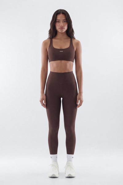 A woman stands facing forward in a studio wearing a matching dark brown activewear set consisting of a sports bra and high-waisted leggings, paired with white socks and sneakers.