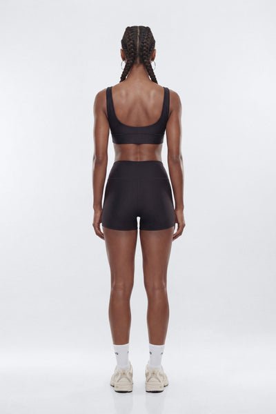 A woman viewed from the back wearing a matching black activewear set consisting of a sports bra and high-waisted shorts, paired with white socks and beige sneakers, against a light gray studio background.