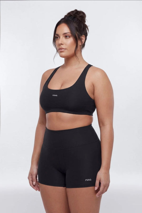 A woman modeling a matching black activewear set consisting of a scoop-neck sports bra and high-waisted athletic shorts, both featuring a small white &