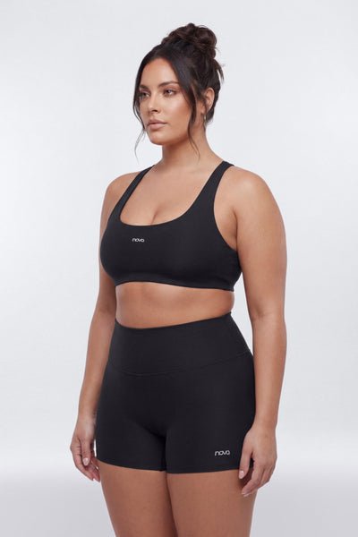 A woman modeling a matching black activewear set consisting of a scoop-neck sports bra and high-waisted athletic shorts, both featuring a small white &