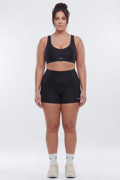 A woman stands against a light gray studio background wearing a matching black activewear set consisting of a scoop-neck sports bra and high-waisted athletic shorts, paired with white socks and beige sneakers.