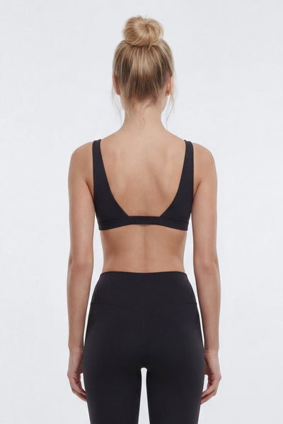 A woman viewed from the back wearing a black sports bra and black leggings against a plain light gray studio background.