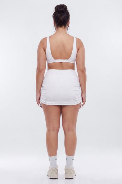 A woman viewed from behind wearing a white two-piece set consisting of a scoop-neck bralette and a ruched mini skirt, white studio background.