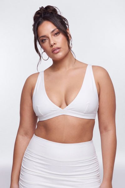 A woman modeling a white ribbed bralette with a crossover front and deep V-neckline, paired with a matching white high-waisted ruched mini skirt, against a light gray studio background.