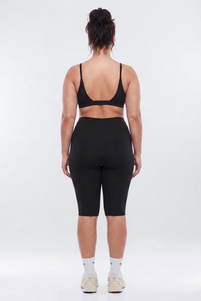 A woman viewed from behind wearing a black sports bra and black fitted biker shorts, standing against a light gray studio background.