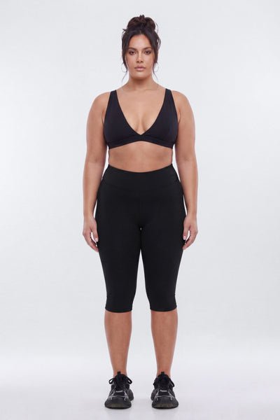 A woman modeling a black activewear set consisting of a scoop-neck crop top and high-waisted capri leggings, paired with black athletic sneakers, against a plain light gray studio background.