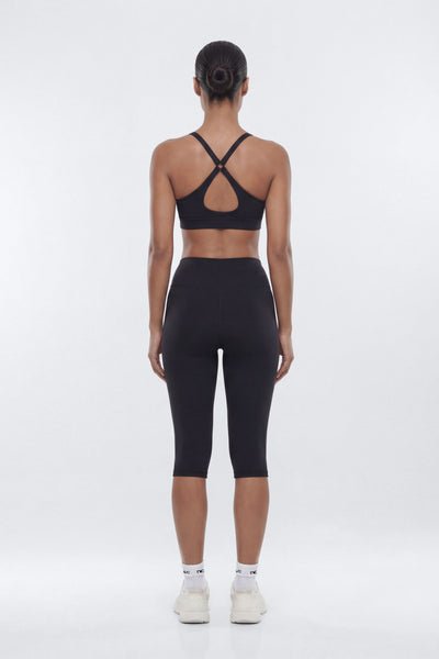A woman viewed from the back wearing a black sports bra with crisscross straps and black capri leggings, standing against a light gray studio background.