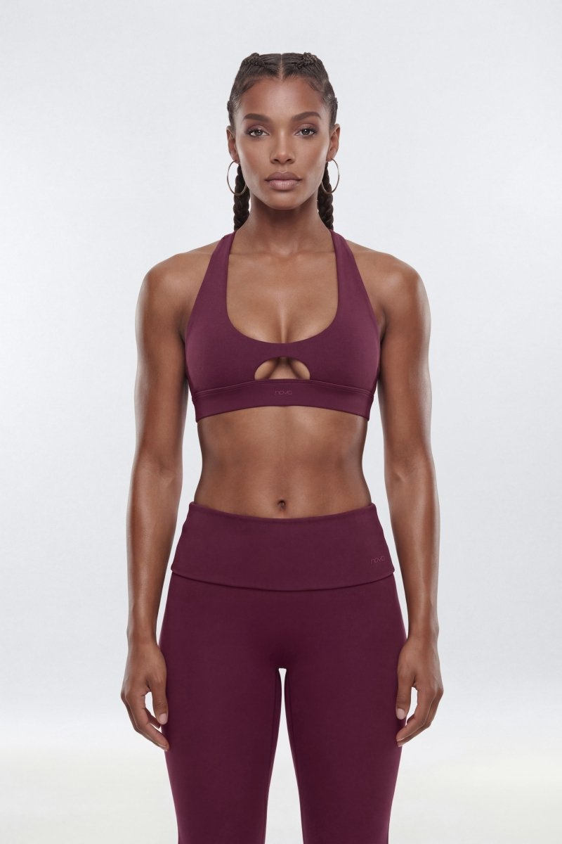 A woman modeling a matching athletic set consisting of a deep plum purple sports bra with a keyhole cutout and high-waisted leggings, shot against a seamless light gray studio background.