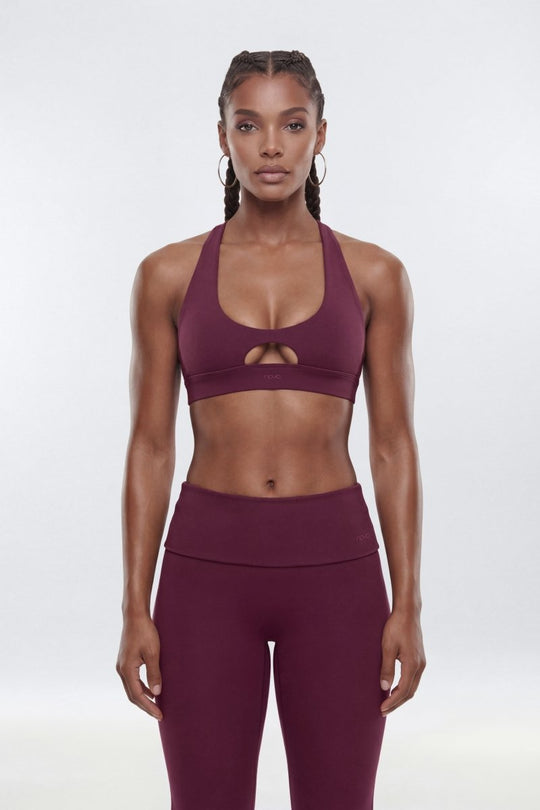 A woman modeling a matching athletic set consisting of a deep plum purple sports bra with a keyhole cutout and high-waisted leggings, shot against a seamless light gray studio background.