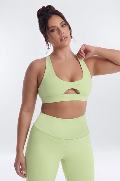 A woman modeling a matching light lime green activewear set consisting of a sports bra with a chest cutout and high-waisted leggings, against a plain light gray studio background.