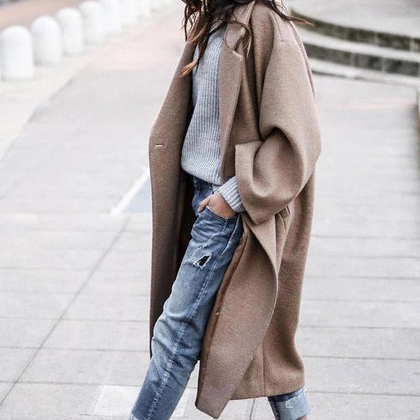 Person wearing a long brown coat over a light blue sweater and jeans on a blurred urban background