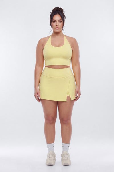 A woman modeling a matching pale yellow athletic set consisting of a halter crop top and a high-waisted wrap mini skirt, paired with white socks and beige sneakers, against a light gray studio background.