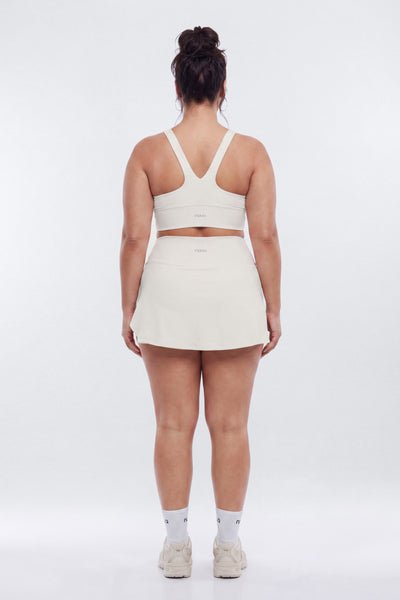 A woman is shown from behind wearing a matching off-white athletic set consisting of a sports bra and a tennis skirt, paired with white socks and sneakers, against a plain white studio background.