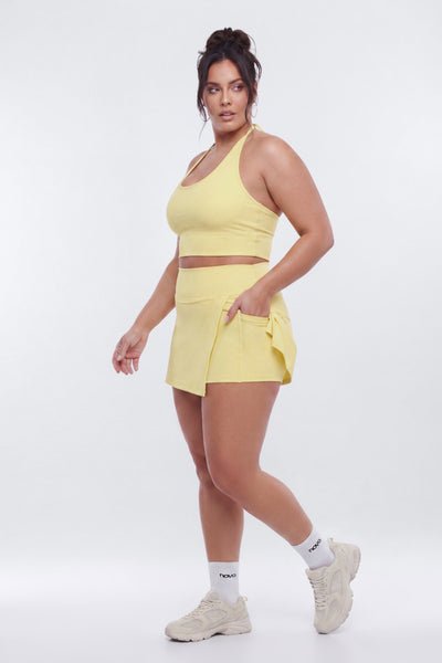 A woman modeling a light yellow athletic set consisting of a halter crop top and a matching wrap-style mini skirt, paired with white socks and beige sneakers, against a plain white studio background.