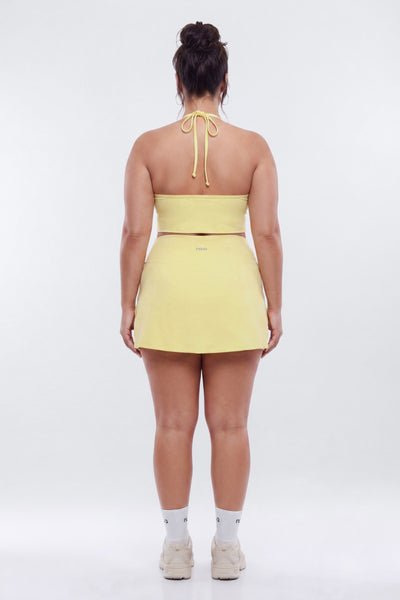 A woman viewed from behind wearing a pale yellow halter mini dress with a tie-back strap, paired with white socks and beige sneakers, against a plain white studio background.