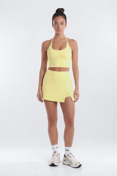 A woman modeling a matching athletic set consisting of a pale yellow sports bra and a high-waisted skort with a side zipper, paired with white socks and sneakers, against a light gray studio background.