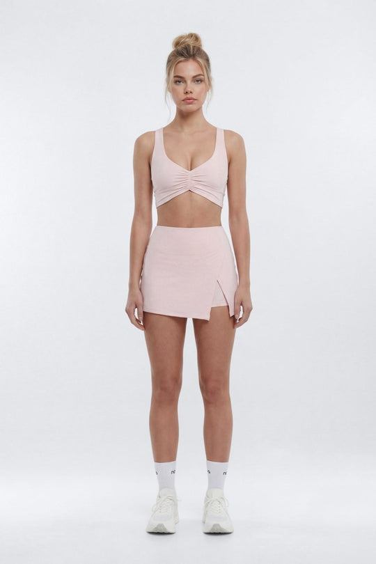 A model stands against a light gray studio background wearing a light pink activewear set consisting of a sports bra with a front twist detail and a matching high-waisted mini skirt with a side zipper.