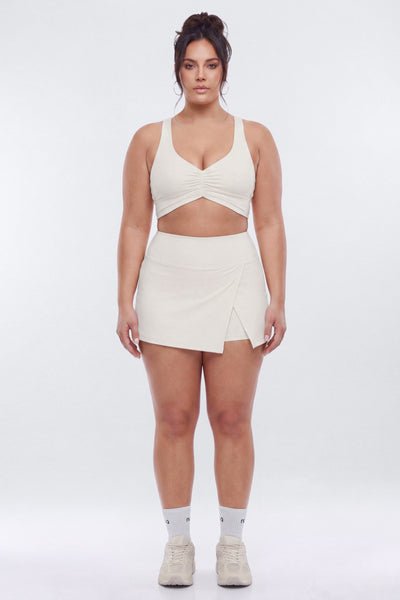 A plus-size model stands against a light gray studio background wearing a matching cream-colored two-piece activewear set consisting of a crossover sports bra and a wrap-style mini skirt, paired with white socks and beige sneakers.