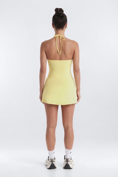 A woman viewed from the back wearing a pale yellow halter mini dress with a tie at the back of the neck, paired with white socks and sneakers, against a light gray studio background.