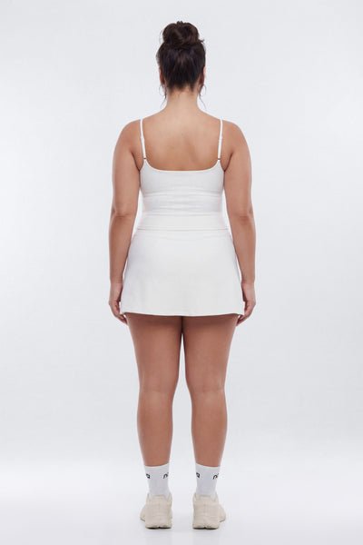 A woman viewed from the back wearing a white sleeveless mini dress with thin straps and a fitted waist, paired with white socks and white sneakers, against a plain light gray studio background.