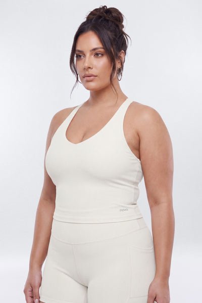 A woman modeling a matching cream-colored activewear set, featuring a racerback tank top and high-waisted shorts, against a plain light gray studio background.