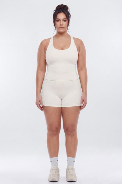 A woman modeling a matching beige ribbed tank top and shorts set, paired with white socks and beige sneakers, against a plain light gray studio background.