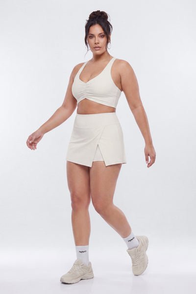 A woman modeling a matching cream-colored activewear set consisting of a wrap-style sports bra and a wrap-style mini skirt, paired with white socks and beige sneakers, against a plain white studio background.