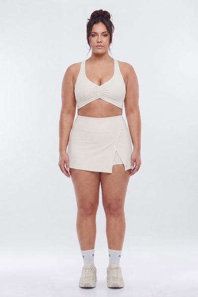 A woman modeling a matching off-white athletic set consisting of a crossover sports bra and a wrap-style mini skirt, paired with white socks and sneakers, against a light gray studio background.