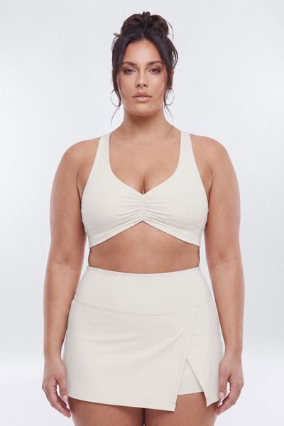 A woman modeling a matching two-piece activewear set in cream, featuring a ruched sports bra and a high-waisted wrap mini skirt, set against a plain light gray studio background.