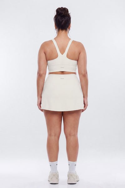 A woman viewed from behind wearing a matching off-white athletic set consisting of a sports bra and a short tennis skirt, paired with white socks and sneakers, against a light gray studio background.
