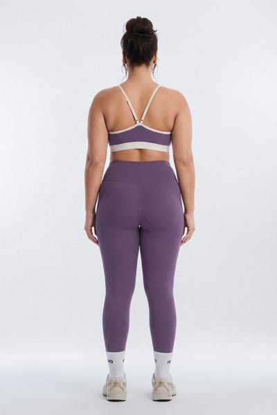 A woman viewed from the back wearing a matching purple activewear set consisting of a sports bra and high-waisted leggings, paired with white socks and beige sneakers, against a light gray studio background.