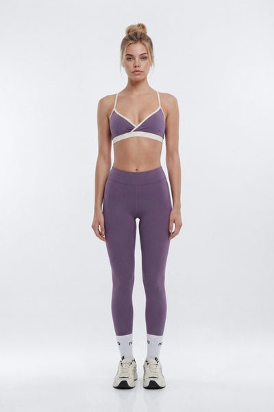 A woman stands facing forward in a studio wearing a matching purple activewear set consisting of a sports bra with white trim and high-waisted leggings, paired with white socks and white sneakers.